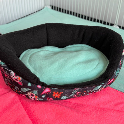 The Curious Catrina Fleece Bed beckons with bewitching charm—black plush fleece adorned in vivid sugar skulls and gothic florals dances across its surface. Nestled atop layers of bright pink and turquoise bedding inside a crisp white wire enclosure, this cozy nest invites your guinea pig to lounge like a little creature of the night. Perfect for spooky-cute pets with a flair for dramatic naps.