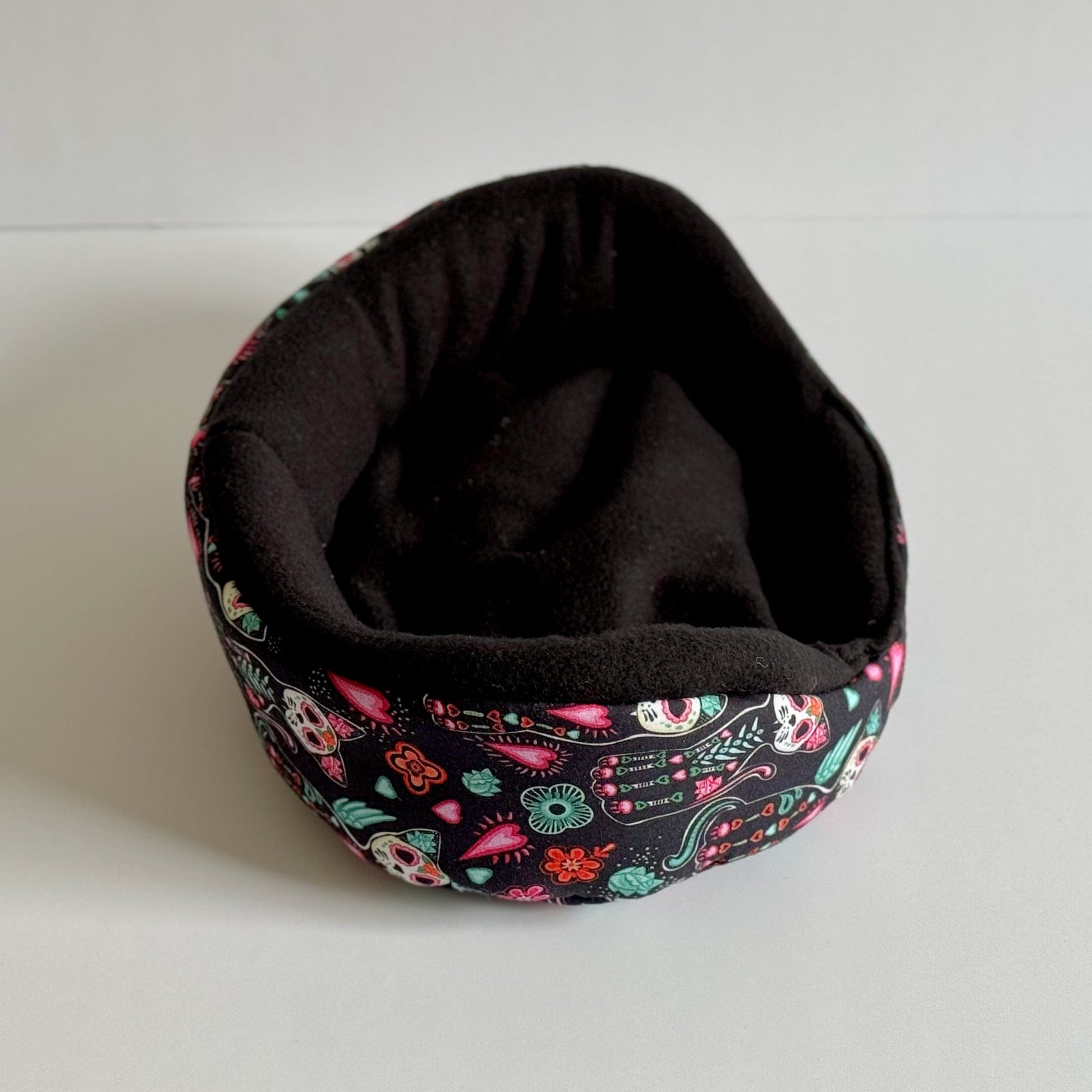 The Curious Catrina Fleece Bed—a whimsically gothic snuggle spot for guinea pigs—features a plush, midnight-black interior wrapped in a bewitching exterior. The oval-shaped bed enchants with vivid sugar skulls, moonlit flowers, and ornate guitars dancing across a shadowy fabric. Perfect for pint-sized familiars seeking gothic-glam coziness, the bed is pictured resting atop a pale surface, ready to treat your small pet to sweetly spooky dreams.