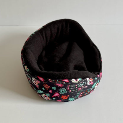 The Curious Catrina Fleece Bed—a whimsically gothic snuggle spot for guinea pigs—features a plush, midnight-black interior wrapped in a bewitching exterior. The oval-shaped bed enchants with vivid sugar skulls, moonlit flowers, and ornate guitars dancing across a shadowy fabric. Perfect for pint-sized familiars seeking gothic-glam coziness, the bed is pictured resting atop a pale surface, ready to treat your small pet to sweetly spooky dreams.