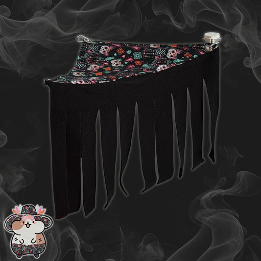 The Curious Catrina Corner Fleece Forest—an enchanting hideaway for small pets—features midnight-black fleece fringe, draping like gothic curtains. Adorned with a vivid, sugar skull pattern and mysterious smoky swirls swirling over a moody dark backdrop. At the bottom left corner, a charming cartoon hamster in a festive sombrero invites your little beastie into this whimsically spooky haven.