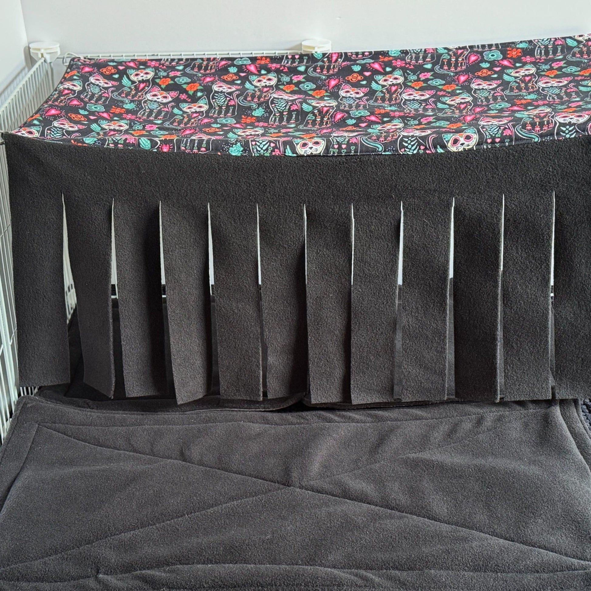 A cozy small pet enclosure is draped in moody, midnight charm—nestled atop a plush, blackened mat, a whimsical Curious Catrina Haybox Fleece Forest curtain tumbles down in gothic slits. Above, playful sugar skull cats lounge amid heart blooms and vibrant blossoms, their candy hues popping against the mystic black backdrop. Perfect for pampered guinea pigs or adventurous little critters who crave a dash of spooky-cute glamour in their hideaways.