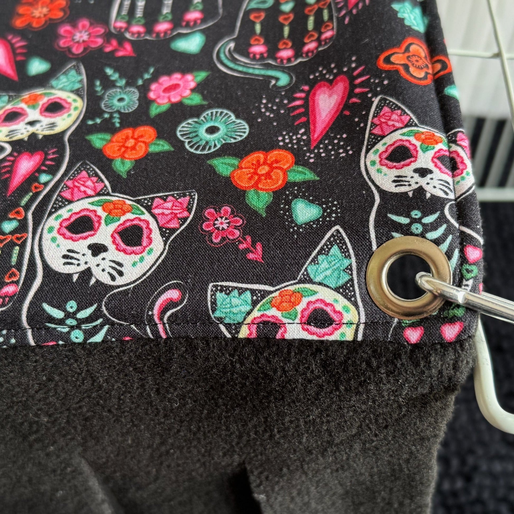 Bewitching close-up of inky black fleece, adorned with vibrant sugar skull kitties, dreamy florals, and bright candy hearts—each design swirling with gothic glam and playful charm. Soft fleece fringe cascades from a shiny silver metal ring, inviting small pets to frolic through this mystical Curious Catrina Haybox Fleece Forest accessory. Perfect for your guinea pig or tiny familiar’s lair!.
