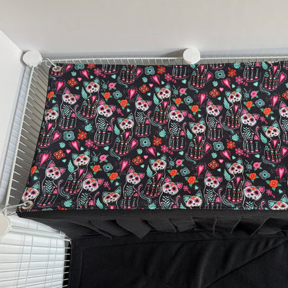 The Curious Catrina Haybox Fleece Forest is a delightfully eerie, soft rectangular fleece blanket adorned with enchanting sugar skull cats and vibrant gothic florals swirling across a midnight-black background. This cozy creation invites your small pet to burrow beneath charmingly spooky patterns, lending a dash of gothic-glam allure and snuggly comfort to any wire enclosure.