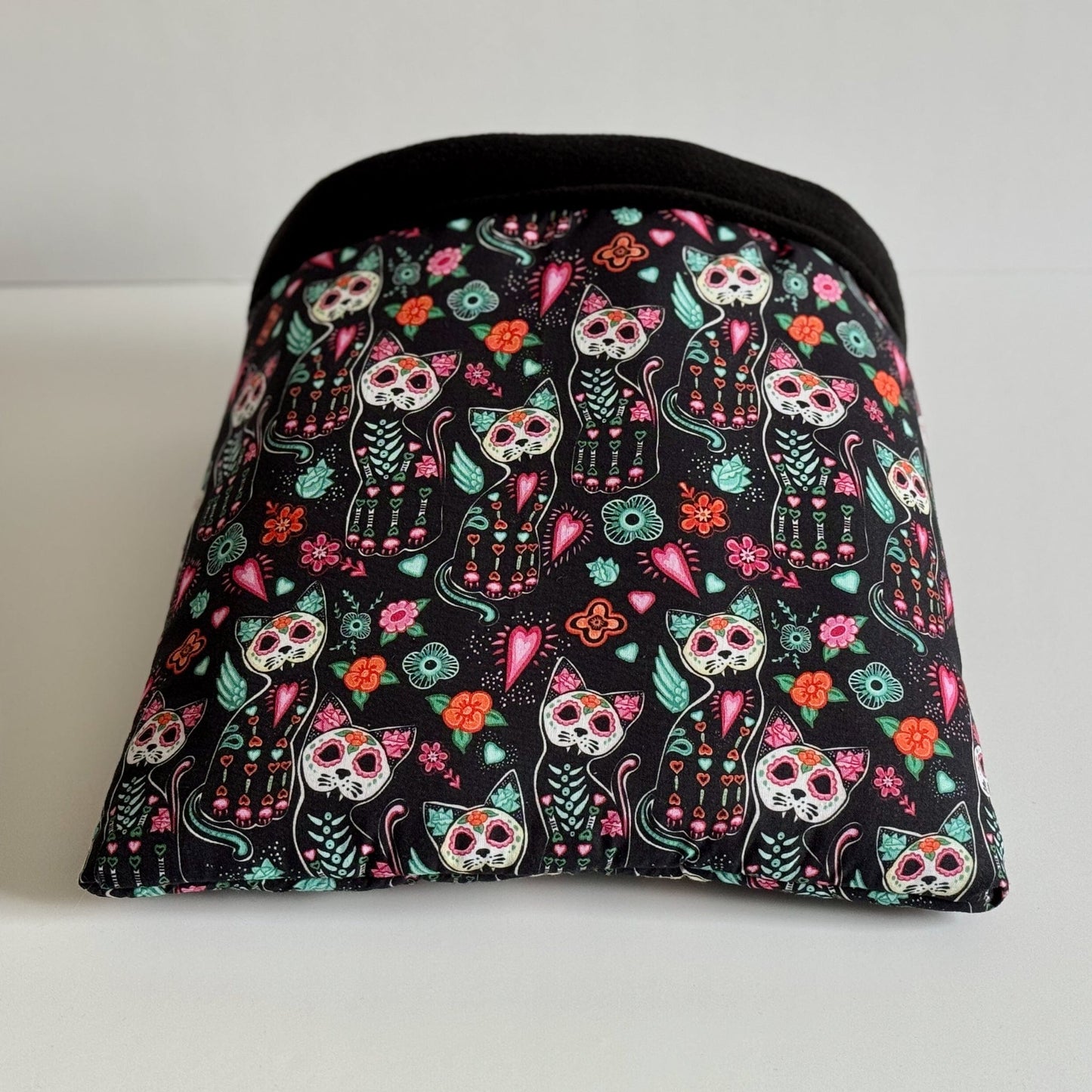 Enchant your little familiar with the Curious Catrina Snuggle Sack—an irresistibly cozy pouch crafted in gothic-glam style. Bewitching midnight-black fabric is adorned with whimsical Day of the Dead skeleton kitties, radiant flowers, and charming heart motifs in vivid candy colors. The plush black fleece lining invites guinea pigs or other small companions to burrow deep into mysterious comfort. Finished with a sleek black trim for extra drama—perfect for cuddles and midnight mischief!.