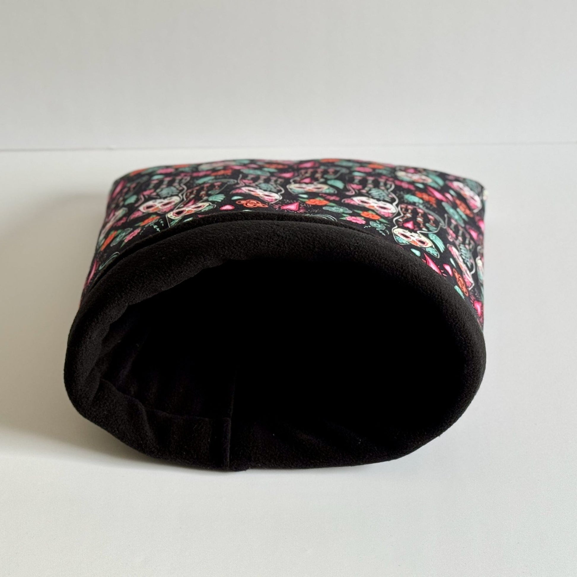 The Curious Catrina Snuggle Sack—a gothic-glam haven for small pets—boasts plush black fleece adorned with enchanting, jewel-bright blooms and whimsical animal skull motifs. Its velvety tunnel shape beckons curious guinea pigs to snuggle in style, like tiny phantoms at a masquerade. Shown resting on a bone-white backdrop, this spooky-cute accessory turns every nap into a moonlit slumber party.