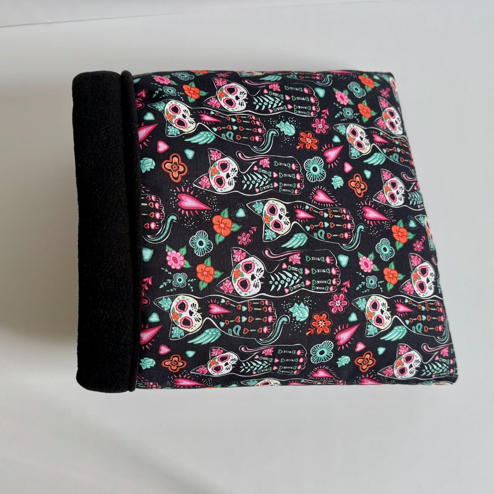 The Curious Catrina Snuggle Sack is an enchanting hideaway for small pets—crafted from plush black fleece and cloaked in a spellbinding print of vibrant Day of the Dead sugar skull cats dancing across an inky background. Its cozy, cave-like shape invites guinea pigs to burrow within, while a sleek raven-black spine adds gothic flair. Perfect for spooky-cute snuggles on chilly nights!.