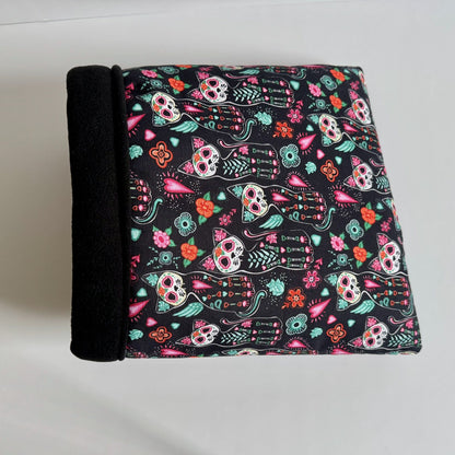The Curious Catrina Snuggle Sack is an enchanting hideaway for small pets—crafted from plush black fleece and cloaked in a spellbinding print of vibrant Day of the Dead sugar skull cats dancing across an inky background. Its cozy, cave-like shape invites guinea pigs to burrow within, while a sleek raven-black spine adds gothic flair. Perfect for spooky-cute snuggles on chilly nights!.