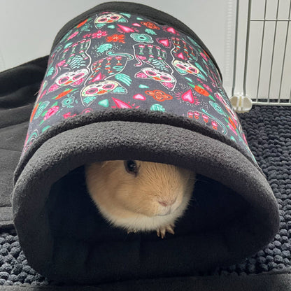 A charming light brown guinea pig gazes out from inside the Curious Catrina Fleece Tunnel—a cozy gothic hideaway adorned with vibrant sugar skull cats in jewel tones, swirling roses, and mystical filigree. The plush tunnel sits atop a velvet-black mat with a cobweb texture, casting shadowy contrast near a white pet cage. Spooky-cute comfort for your small companion’s nightly haunt!.