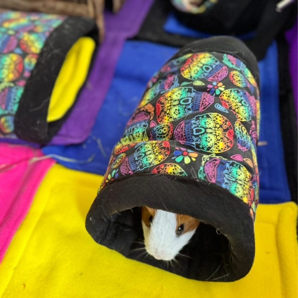 A guinea pig peers from a sugar skull fleece tunnel on a golden mat, surrounded by plush sacks—spooky-cute Frightful Fiesta fun!.