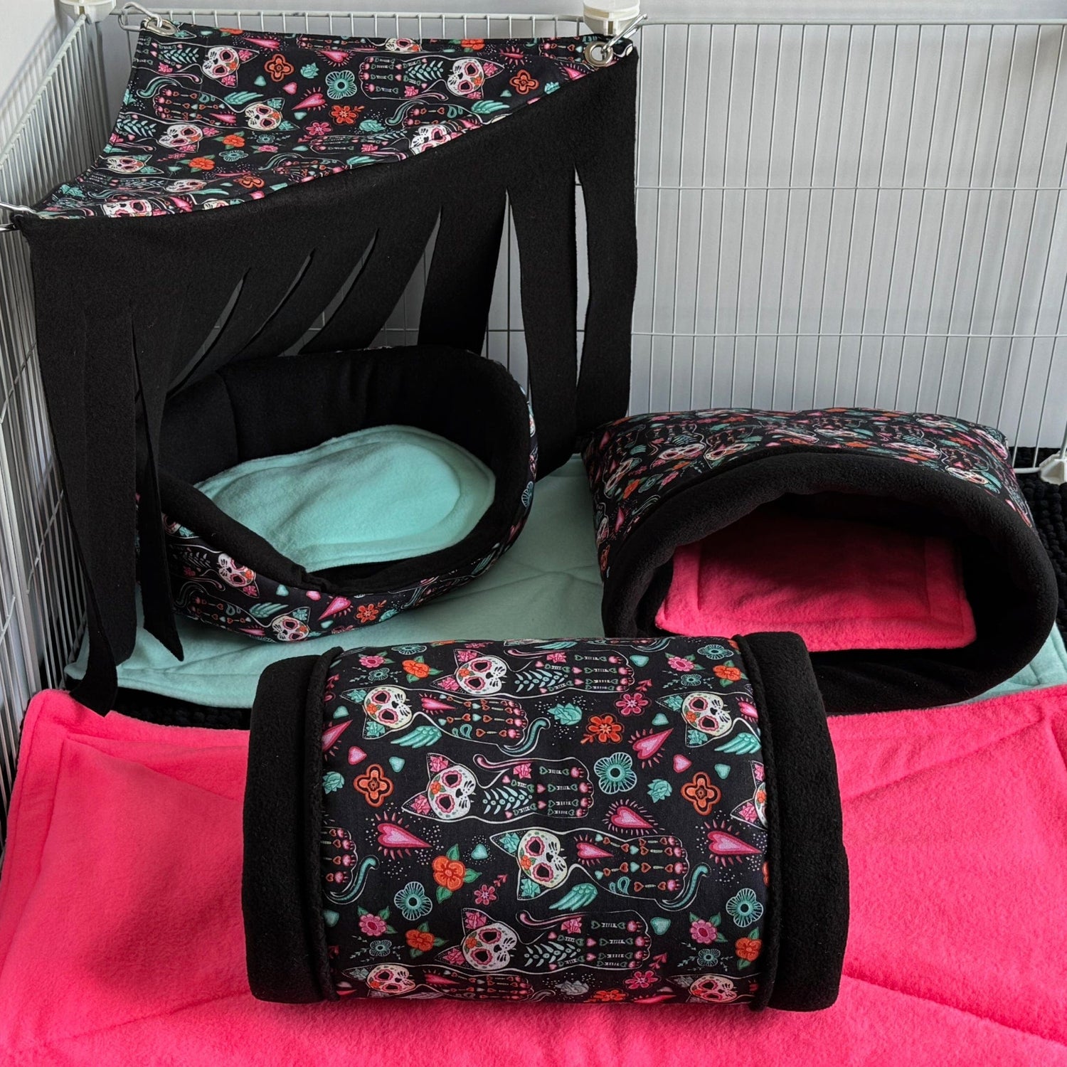 A bewitching set of small animal beds and tunnels adorned with enchanting sugar skulls and whimsical florals, nestled inside a wire enclosure. The gothic-glam bedding collection includes a dramatic canopy with flowing black fabric, a cozy tunnel and mysterious cave hideaway, plus plush mats—all decked out in midnight black with pops of teal and pink for spellbinding flair. Perfect for pampered guinea pigs or other tiny familiars seeking comfort in spooky-cute style.