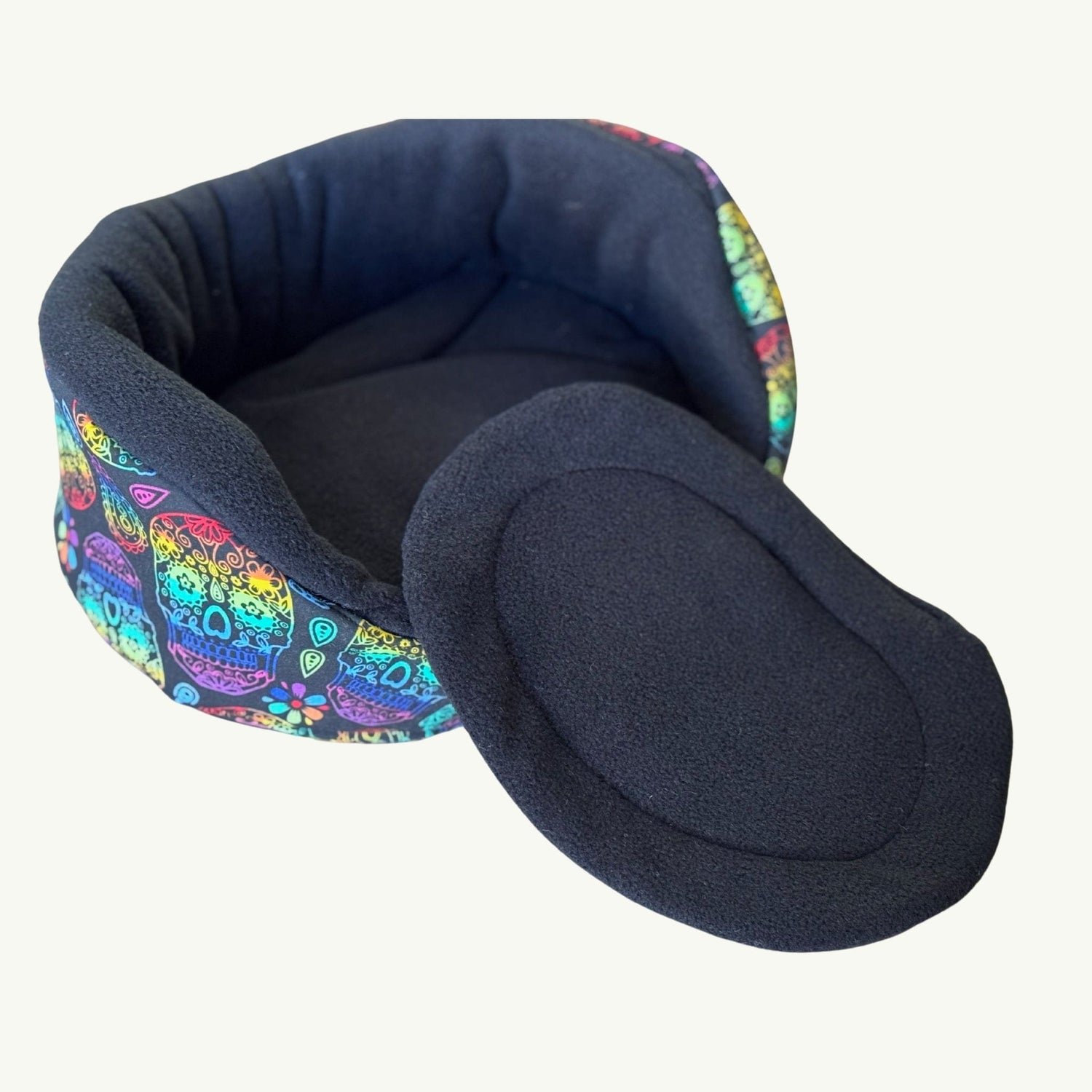 A moonlit-black, round fabric pet bed adorned with enchanting sugar skulls in pops of pink, teal, and gold dancing along its outer sides. In front lies a matching cushion—plush and inviting—ready for your small familiar’s most bewitching dreams. Both bed and cushion rest upon a plain white background, making their gothic-glam details pop like midnight magic.