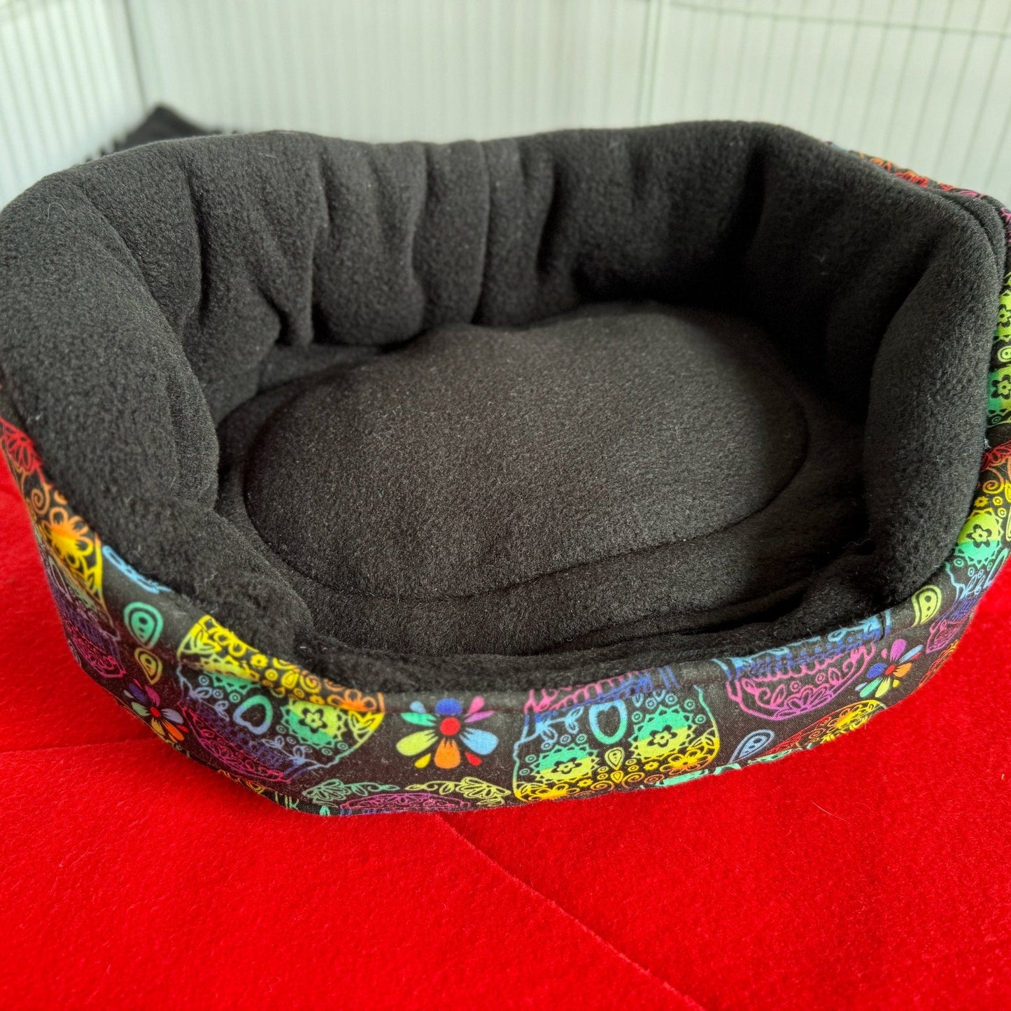 The Frightful Fiesta Fleece Bed beckons your tiny familiar into dreamy darkness—a plush, round nest lined in midnight-black fleece, crowned with a merry riot of gothic-glam blossoms and festive flourishes in electric neon. Perfect for guinea pigs or small familiars who crave comfort with a dash of spooky celebration, shown atop a vibrant red stage that highlights every enchanting detail.