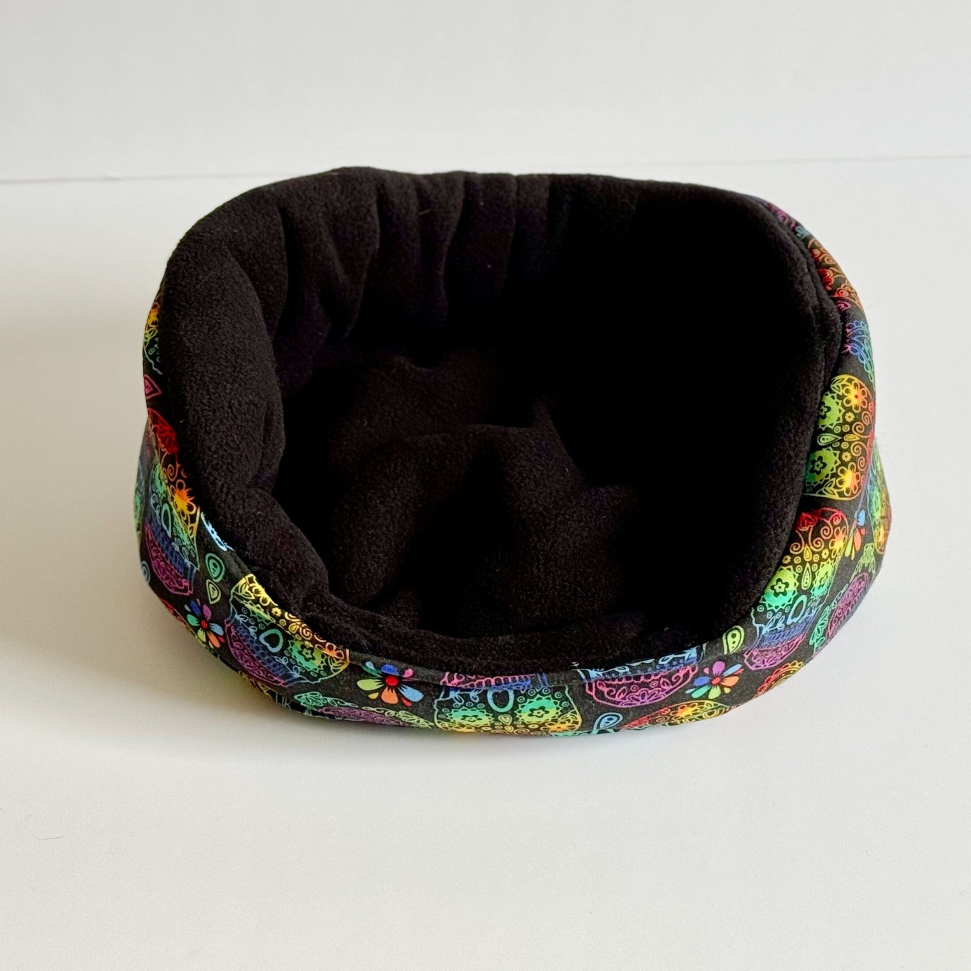 The Frightful Fiesta Fleece Bed awaits—a bewitchingly cozy nest for small pets! This enchanting black fleece bed features a vibrant, gothic-glam rim adorned with playful cartoon cat faces and whimsical flowers in midnight hues and pops of jewel tones. Perfectly sized for guinea pigs to snuggle into a mysterious moonlit slumber. Shown atop a stark white background for spellbinding contrast.
