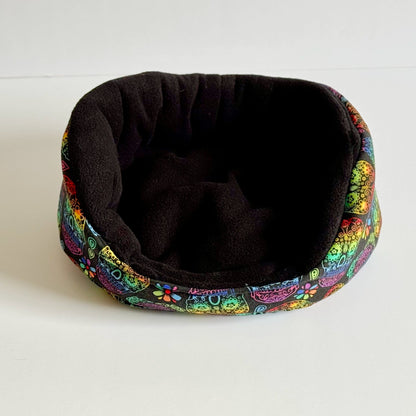 The Frightful Fiesta Fleece Bed awaits—a bewitchingly cozy nest for small pets! This enchanting black fleece bed features a vibrant, gothic-glam rim adorned with playful cartoon cat faces and whimsical flowers in midnight hues and pops of jewel tones. Perfectly sized for guinea pigs to snuggle into a mysterious moonlit slumber. Shown atop a stark white background for spellbinding contrast.