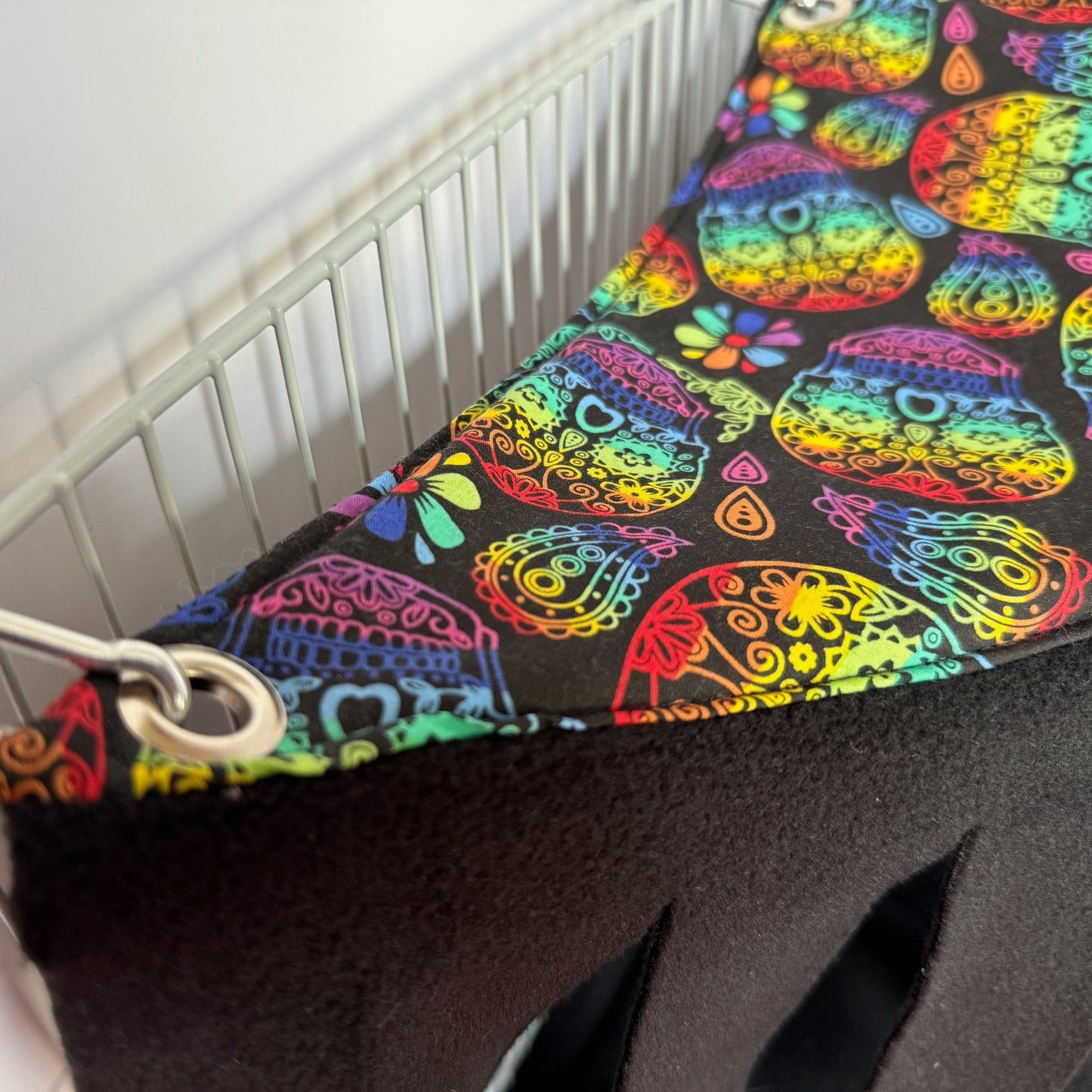 A close-up of the Frightful Fiesta Corner Fleece Forest, a whimsically gothic hideaway for guinea pigs and small pets. This handmade hammock dazzles with vibrant sugar skulls and enchanting florals atop inky black fleece, conjuring festive yet mysterious vibes. Snuggly soft lining promises cozy midnight naps, while sturdy metal grommets and clips fasten it neatly to a white wire cage—a bewitching nook for your little familiars to frolic or plot their next adventure!.