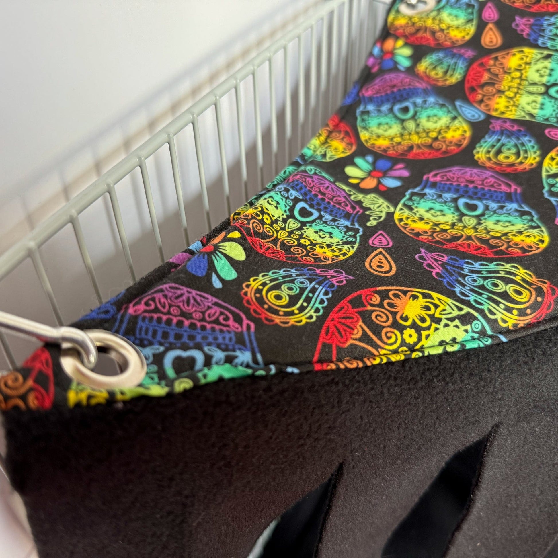 A close-up of the Frightful Fiesta Corner Fleece Forest, a whimsically gothic hideaway for guinea pigs and small pets. This handmade hammock dazzles with vibrant sugar skulls and enchanting florals atop inky black fleece, conjuring festive yet mysterious vibes. Snuggly soft lining promises cozy midnight naps, while sturdy metal grommets and clips fasten it neatly to a white wire cage—a bewitching nook for your little familiars to frolic or plot their next adventure!.