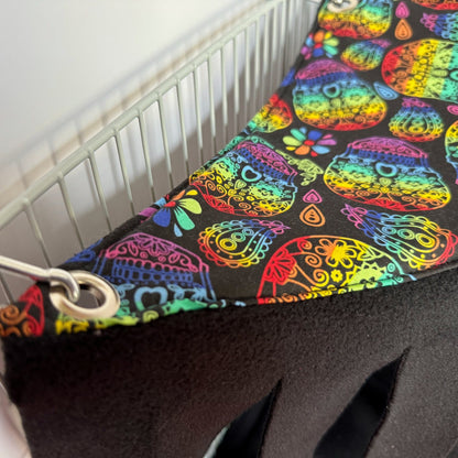 A close-up of the Frightful Fiesta Corner Fleece Forest, a whimsically gothic hideaway for guinea pigs and small pets. This handmade hammock dazzles with vibrant sugar skulls and enchanting florals atop inky black fleece, conjuring festive yet mysterious vibes. Snuggly soft lining promises cozy midnight naps, while sturdy metal grommets and clips fasten it neatly to a white wire cage—a bewitching nook for your little familiars to frolic or plot their next adventure!.