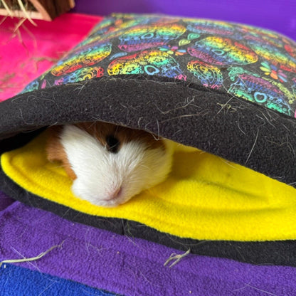 A curious guinea pig peeks out from a plush fleece tunnel, bewitched with whimsical owls in moonlit hues. The tunnel perches atop our Frightful Fiesta Matching Pee Pads—ghoulishly cheerful in haunted-house brights and sunbeam yellow lining—banishing messes like magic. All rests upon sumptuous purple and midnight blue blankets, creating a lair both spooky and sweet for your pampered pet.