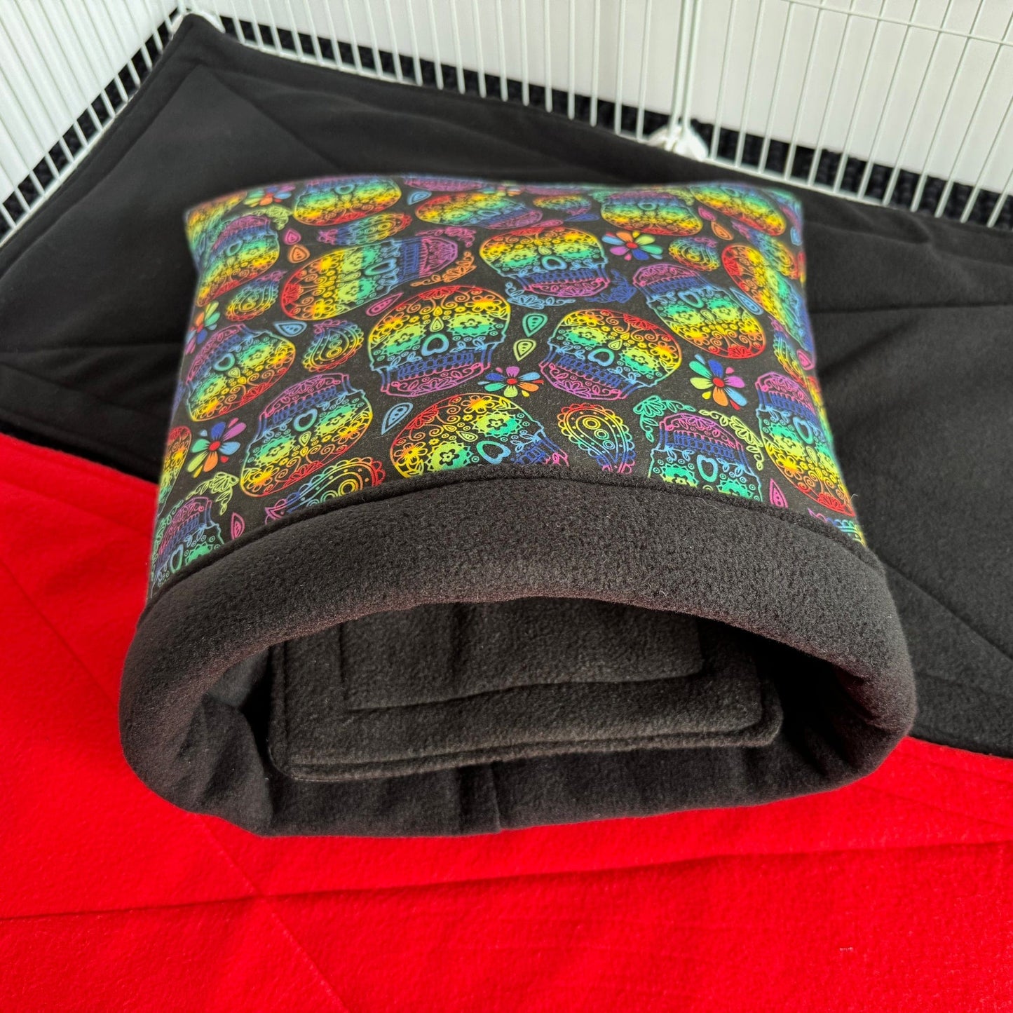 The Frightful Fiesta Snuggle Sack enchants with vivid sugar skulls and enchanting florals swirling across midnight-black fabric, lined inside with sumptuous black fleece—an irresistibly cozy throne for guinea pigs or other petite familiars. Nestled atop a dramatic red-and-black mat within a gothic-white wire enclosure, this snuggle sack is equal parts spooky charm and plush comfort—a bewitching hideaway for your tiniest companions.