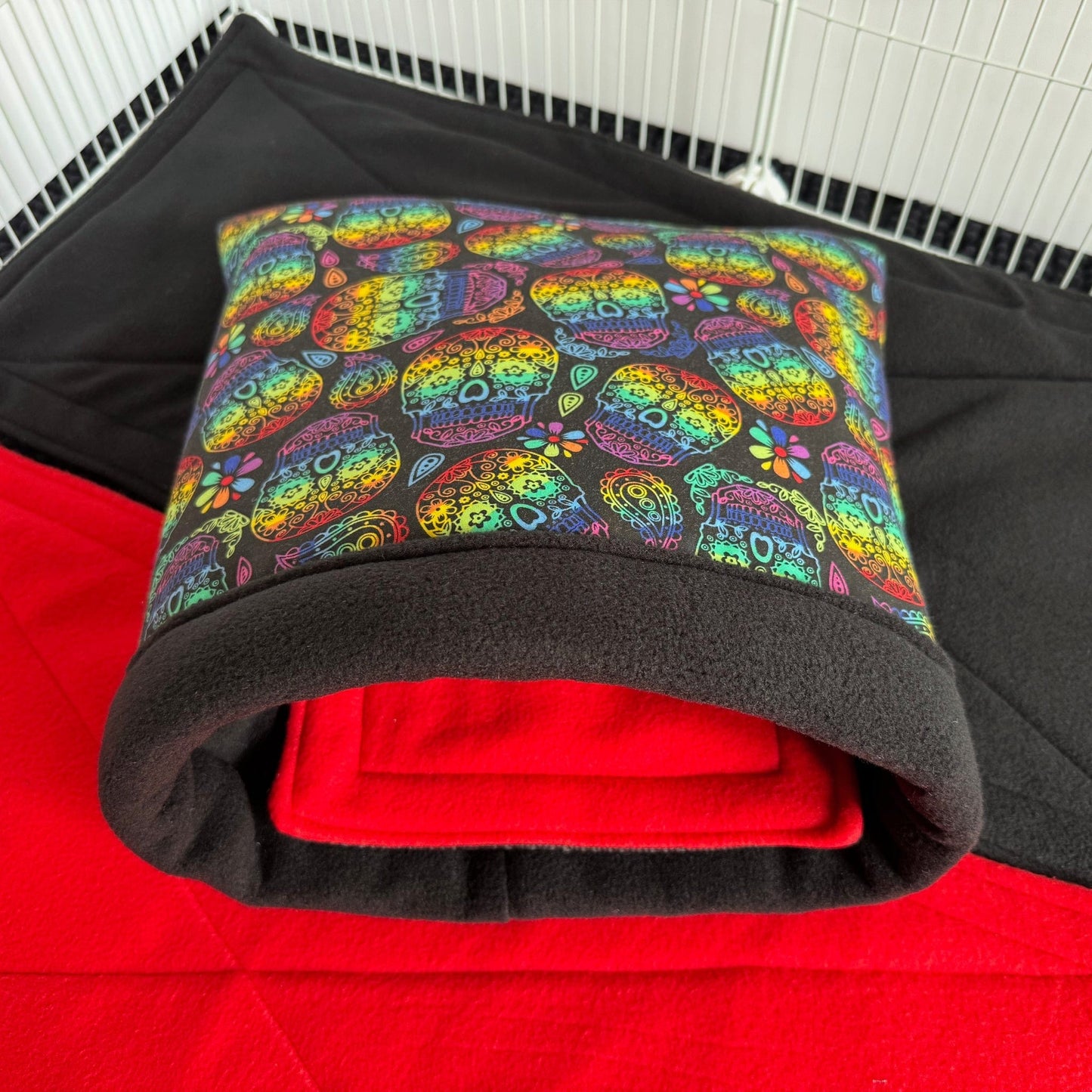 The Frightful Fiesta Snuggle Sack awaits—a whimsical lair of cozy magic for your small companion, draped in vivid sugar skulls and crowned with inky black trim. Plush midnight-soft fleece lines the inside, promising enchantingly snug dreams. Nestled atop layers of crimson and ebony bedding within a wire pet cage, this gothic-glam hideaway is simply spellbinding—perfect for guinea pigs (or other tiny creatures of the night) to snuggle and snooze in style.