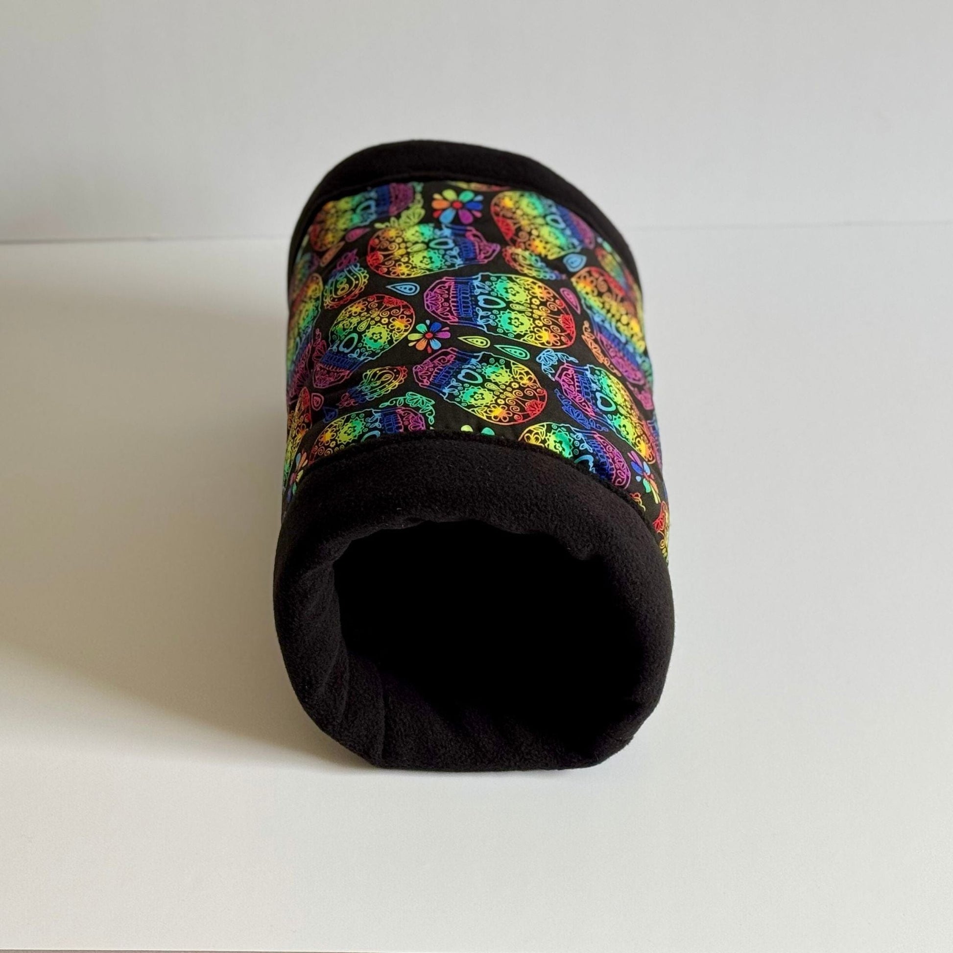 The Frightful Fiesta Fleece Tunnel—a whimsically gothic hideaway for small pets! This cozy, cylindrical tunnel beckons with a vibrant pattern of playful sugar skulls in crisp purples, pinks, and teals, set against midnight-black fleece. Plush and luxuriously lined for burrowing ghouls and glamorous zoomies alike, the tunnel stands upright on a pale background, inviting your furry familiars to snuggle in spooky-cute style. Perfect for guinea pigs or other little dreamers craving a dash of haunted charm!.