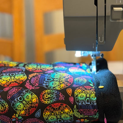 Close-up of a sewing machine weaving magic into the Frightful Fiesta Fleece Tunnel—plush black fabric bedecked with beguiling, candy-bright sugar skulls and gothic florals. The soft, midnight-hued fleece lining promises enchanting snuggles, spirited zoomies, and cozy hideaways for your small pet’s most spine-tingling dreams.