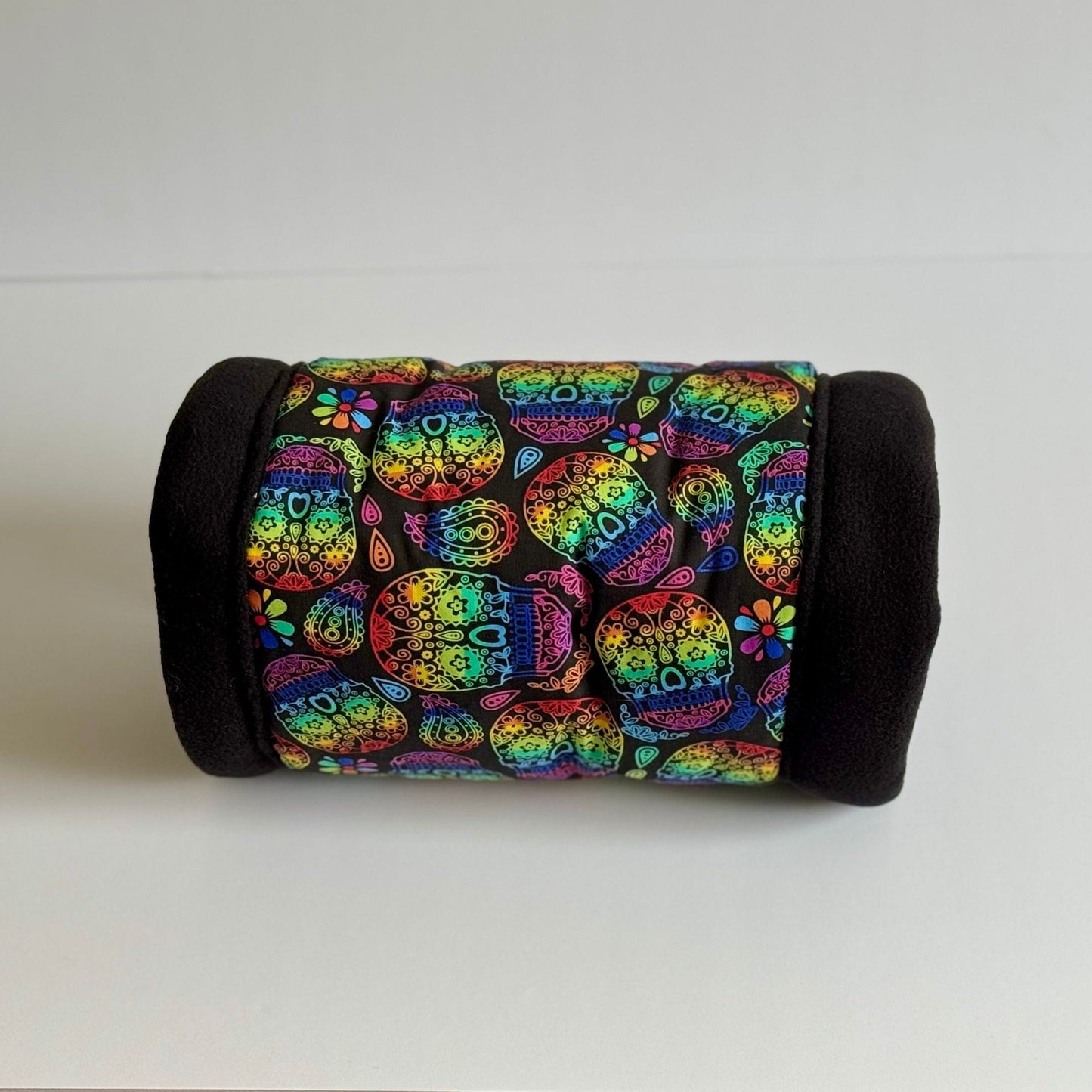 A bewitchingly cozy fleece tunnel for guinea pigs, named the Frightful Fiesta. This washable tunnel features gothic black ends and a fabulously vibrant center adorned with whimsical Day of the Dead sugar skulls and dreamy blooms in pops of teal, pink, yellow, and purple. The plush hideaway sits atop a crisp white background—perfect for pampered pets who love a dash of spooky-cute flair!.
