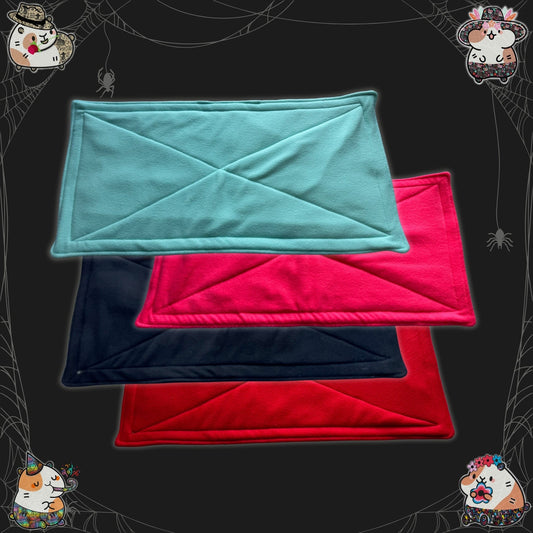 Four snuggly fleece liners and matching pee pads—one each in enchanting teal, bubblegum pink, midnight black, and blood-red—are neatly stacked atop a moody dark backdrop. Cute gothic cartoon critters gaze curiously from the shadows, while playful spider webs and friendly little spiders adorn the corners, spinning cozy magic for your beloved small pets.