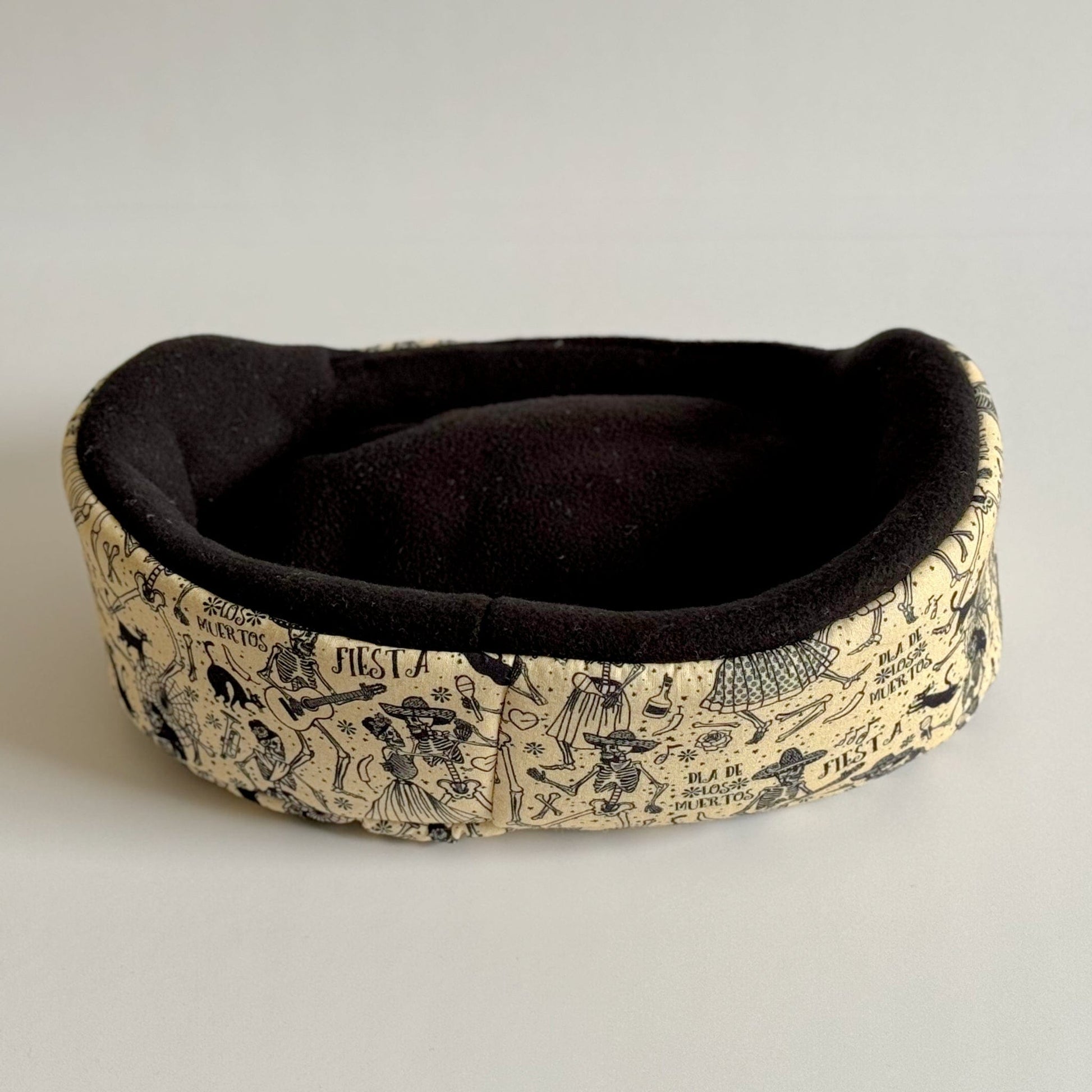 The Tombstone Tango Fleece Bed—a delightfully eerie resting spot for your tiny familiars! This small, oval pet bed features a plush, midnight-black interior where guinea pigs can curl up in cozy darkness. Its pale cream exterior is enchanted with hand-drawn gothic illustrations—think playful tombstones and swirling filigrees—plus mystical Spanish script winding along the sides. Perfect for small pets wanting to snooze in spooky style! Displayed on a simple light backdrop.