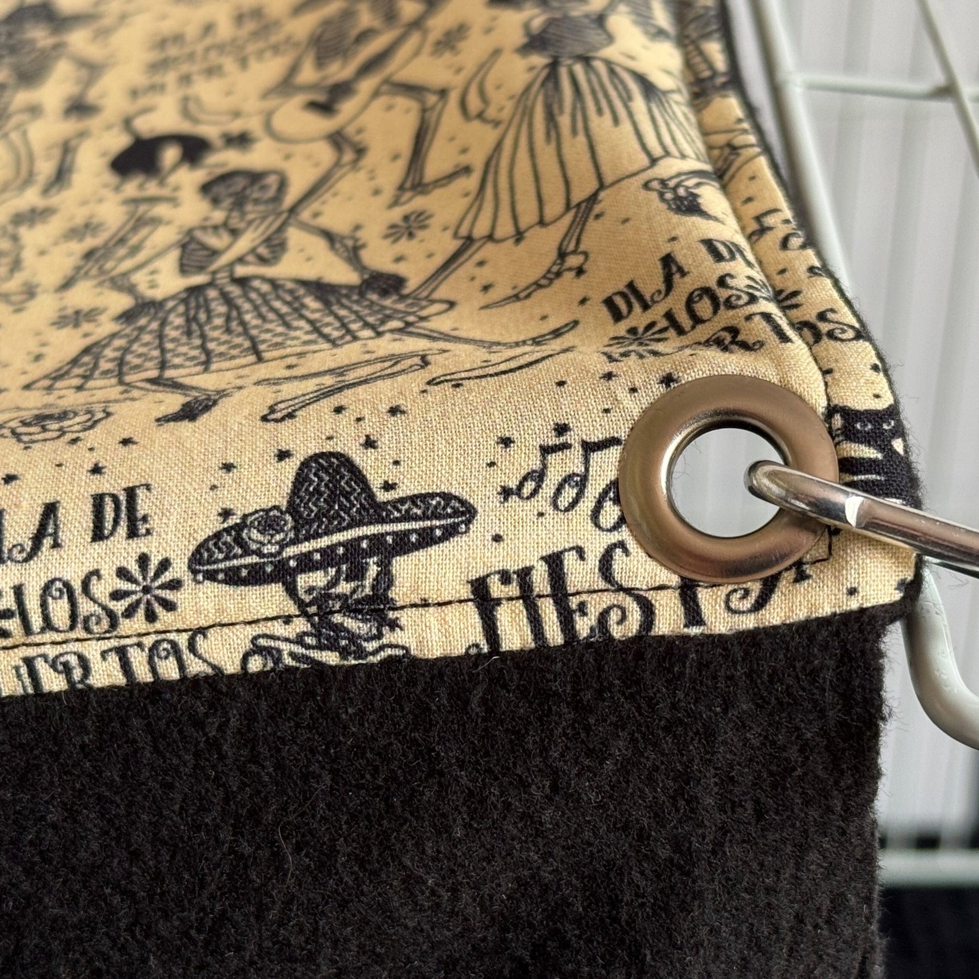 Spooky-cute alt text: Close-up of the Tombstone Tango Large 2x1 Fleece Forest—a dreamy playground for small pets! Wisps of soft fleece in dusky tan and midnight black dangle in bewitching stripes, trimmed with festive Día de los Muertos skulls and swirling Spanish script. Shimmering grommets hold each mystical fringe to a sturdy hook. The plush black forest floor below is perfect for tiny toes to tiptoe through moonlit shadows—cozy, enchanting, and brimming with gothic-glam allure!.