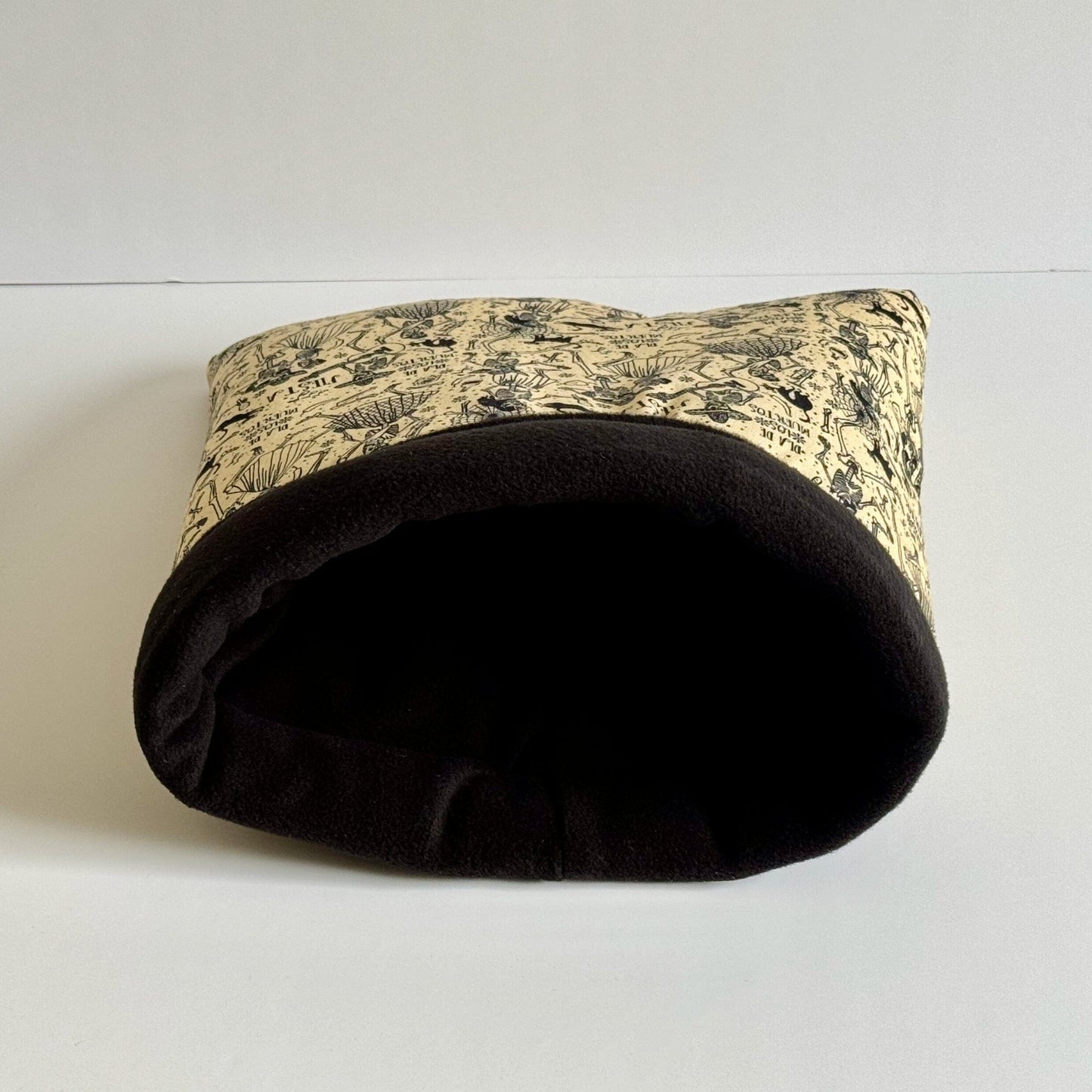 The Tombstone Tango Snuggle Sack—a cozy crypt for your cuddly companion! This gothic-glam pet cave features a midnight-black fleece interior for ultimate snuggle darkness, while the outside gives off haunted elegance with creamy fabric adorned in black vines, bats, and enchanted forest details. Shown resting on its side, mouth agape like a mystical mausoleum door—just waiting to welcome your guinea pig or tiny familiar into luxurious spooky slumber.