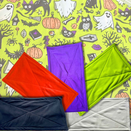 Enchant your cage with five goth-glam fleece pee pads—red, purple, green, black, white—resting on a spooky-cute Halloween haunt.