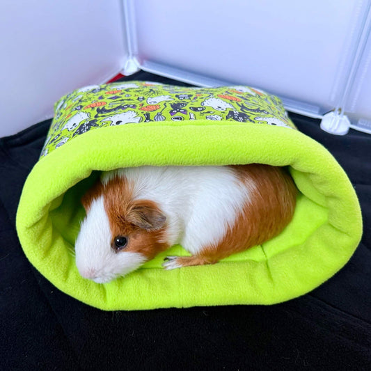 A brown and white guinea pig lounges in a velvet-soft, cartoon-clad snuggle sack—cozy mischief on a midnight-black stage.