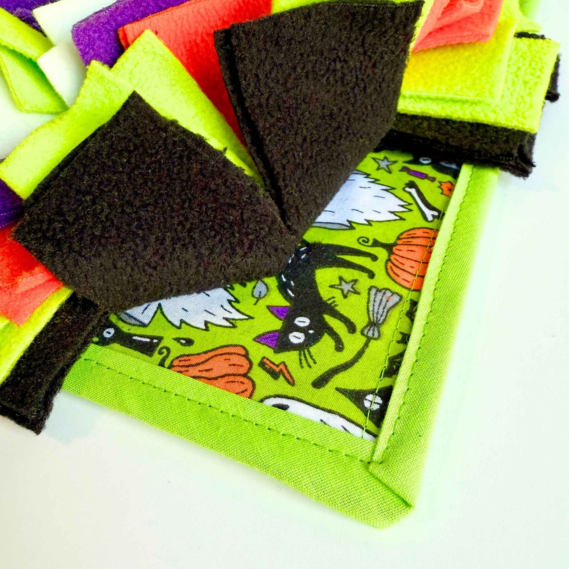 Zoom in on the Trickster’s Treats Snuffle Mat—swirls of black, neon green, and orange felt with gothic cats and pumpkins for spooky-cute foraging.